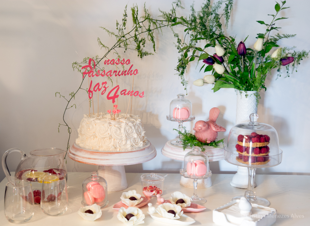 Mesa com bolo branco, decoração floral natural, doces rosa e jarra de chá com limão e framboesas