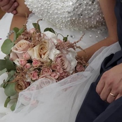Ramo de noiva com flores rosas e creme nas mãos de mulher de vestido branco bordado, ao lado de homem de fato azul
