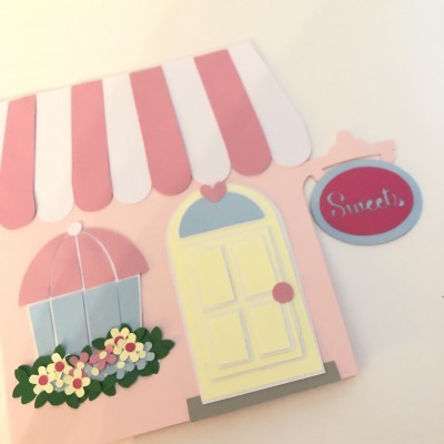 Cartão decorativo em formato de casa com porta amarela, toldo rosa e branco, flores coloridas e letreiro 'Sweets'.