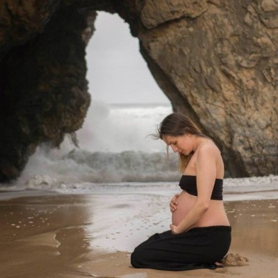 Mulher grávida ajoelhada na praia com mar e rochas ao fundo