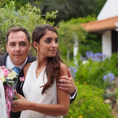 Homem de fato e mulher com vestido branco ao ar livre em frente a casa branca com telhado de telhas