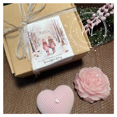 Caixa de presente com laço e etiqueta com ilustração e texto, ao lado duas velas rosas em forma de coração e flor