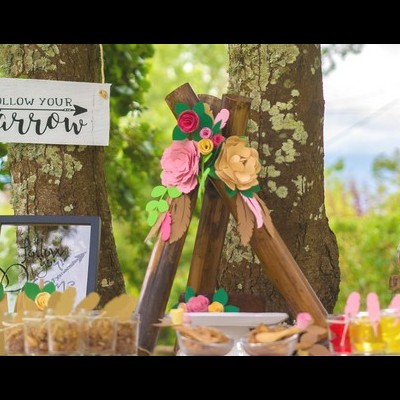 Mesa decorada ao ar livre com placa 'FOLLOW YOUR ARROW' pendurada na árvore e flores artificiais