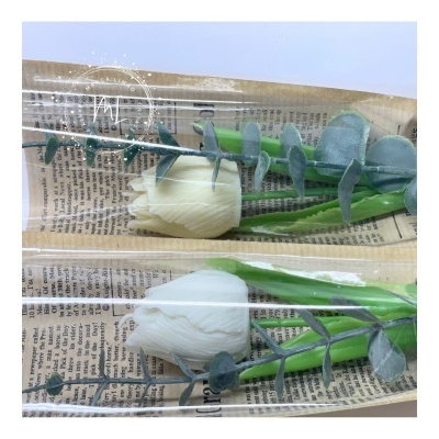 Duas flores artificiais brancas com folhas verdes e ramos azuis embaladas em plástico transparente sobre papel impresso