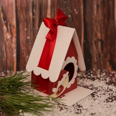 Casa decorativa vermelha e branca com laço vermelho e figura de rena, em cenário de inverno com neve artificial e ramos de pinheiro