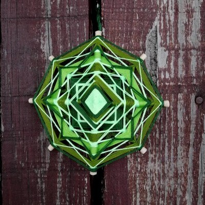 Decoração pendurada hexagonal feita com fios verdes e brancos sobre fundo de madeira vermelha