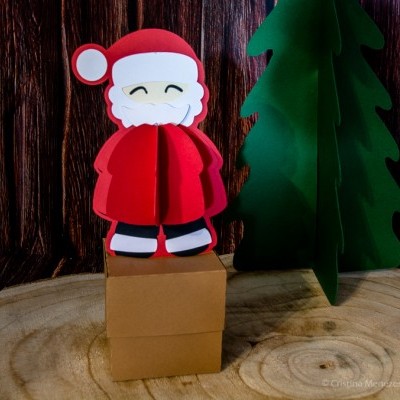 Figura de Pai Natal vermelho e branco em cartão 3D sobre caixa castanha com árvore verde cartesiana e fundo madeira