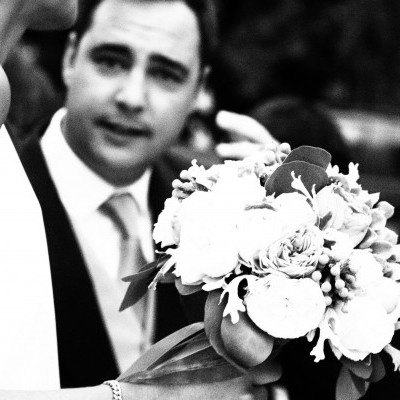 Ramo de flores brancas na mão de uma mulher com vestido branco, homem de fato ao fundo