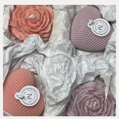 Cinco velas decorativas em forma de coração e flores em tons de rosa e lilás, embaladas com papel branco amassado.