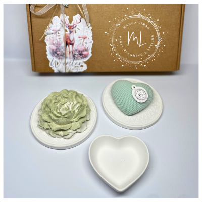 Sabonetes decorativos em forma de flor e coração com suporte branco e caixa de cartão castanho ao fundo.