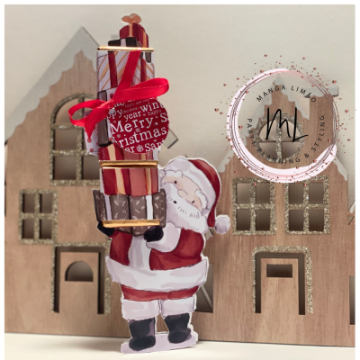 Figura de Pai Natal em madeira com pilha de presentes e casas de madeira ao fundo