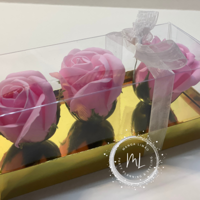 Caixa transparente com três sabonetes em forma de rosa cor-de-rosa e embalagem dourada com laço branco