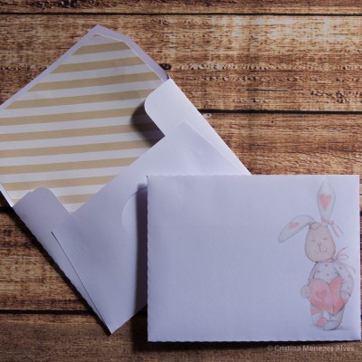 Dois envelopes de papel branco, um aberto com interior dourado às riscas e outro fechado com desenho de coelho cor-de-rosa segurando coração
