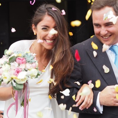 Noiva com vestido branco e ramo de flores e noivo de fato cinzento sorrindo com pétalas caindo