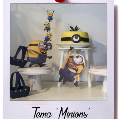 Decoração com bolo e figuras dos Minions em ambiente com mesas brancas e bolsas azuis.