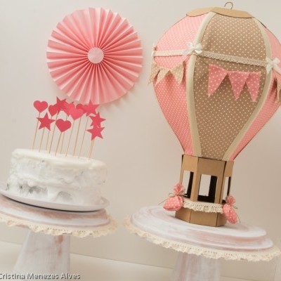 Bolos decorados em suportes brancos com detalhes rendados, um com toppers cor de rosa e outro com balão de ar quente decorativo em papel.