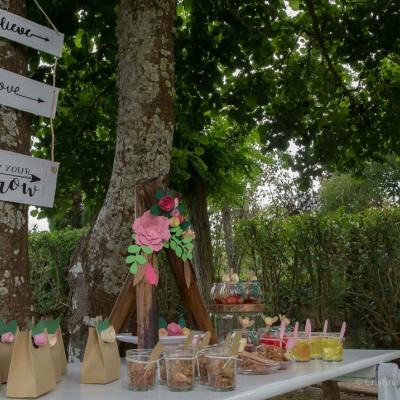 Mesa ao ar livre com pacotes, frascos, copos de sobremesa e painéis de madeira com texto pendurados em árvore