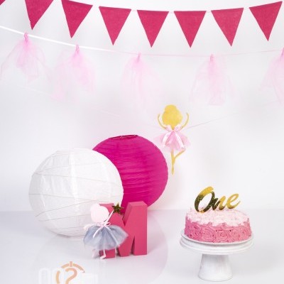 Decoração de festa com bolas de papel, bailarinas, letra M vermelha e bolo rosa decorado com a palavra One.