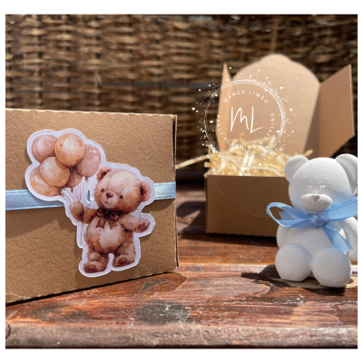 Caixa castanha com etiqueta de urso e balões junto a ursinho branco com fita azul em superfície de madeira