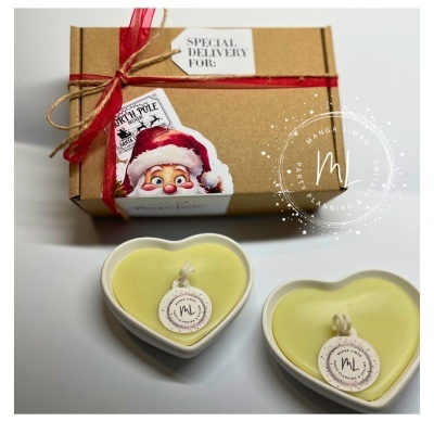 Duas velas amarelas em recipientes corações de cerâmica branca junto a caixa castanha com fita vermelha e etiqueta de Natal.