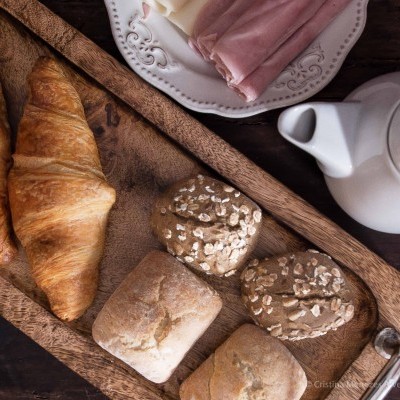 Tabuleiro de madeira com pães variados, prato com fiambre e jarro branco de cerâmica