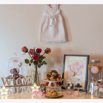 Conjunto decorativo com vestido branco, flores, quadro, bebidas, bolos, letras de madeira e luzes em aparador