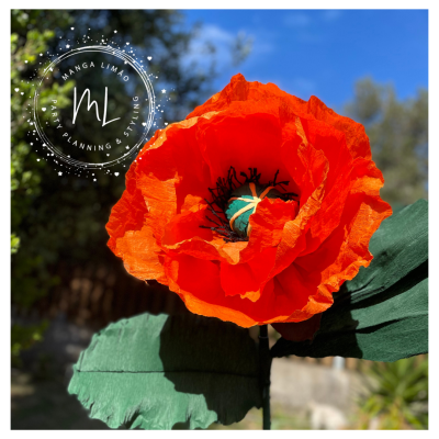 Flor artificial laranja grande com folhas verdes em fundo exterior azul e verde.