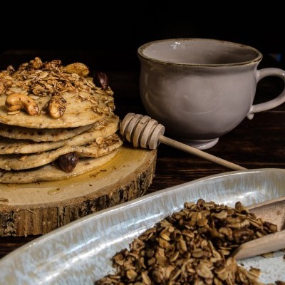 Panquecas com granola e mel em cima de base de madeira, xícara branca e colher de mel numa mesa de madeira