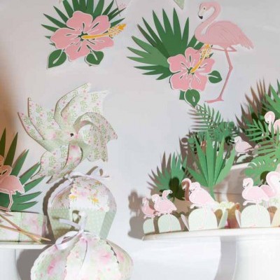 Decoração de festa tropical com flamingos cor-de-rosa, flores, folhas e elementos em papel com padrão floral em fundo branco.