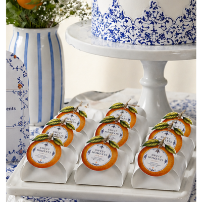 Caixas pequenas brancas para doces com etiquetas decoradas com laranja em base de bolo branca, com decoração azul e vaso com flores e laranja ao fundo.