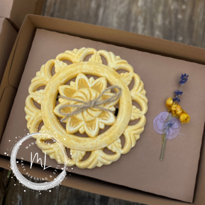 Sabonete decorativo redondo amarelo com padrão floral e flores secas ao lado, numa caixa de cartão marrom