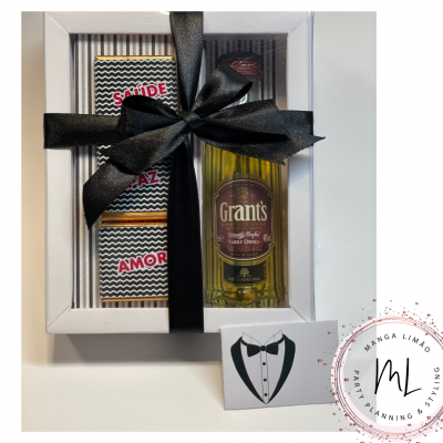 Caixa de presente com mini garrafa de whisky Grant's e três barras de chocolate com mensagem 'SAÚDE PAZ AMOR', decorada com fita preta.
