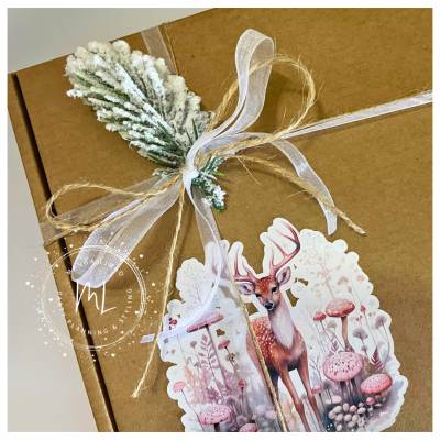 Caixa de presente decorada com ramo neve, fita transparente, corda rústica e etiqueta com veado e cogumelos.
