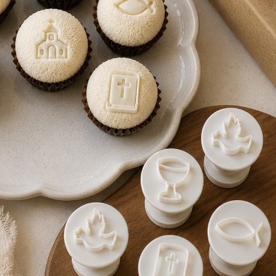 Carimbos brancos com símbolos religiosos e bolos decorados com os mesmos símbolos num prato branco e tábua de madeira ao lado de caixa com laço creme