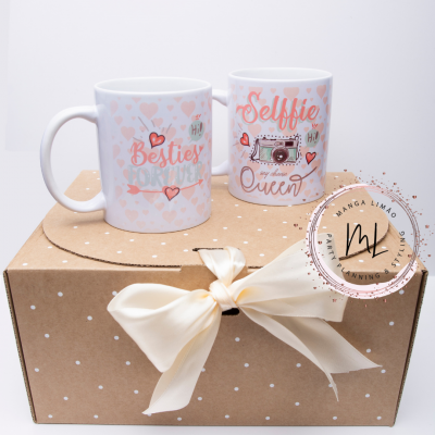 Duas canecas brancas com corações cor-de-rosa e textos, em caixa castanha com bolinhas brancas e laço branco