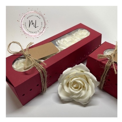 Caixas vermelhas com rosas artificiais brancas dentro, laço de corda e logo ao fundo.