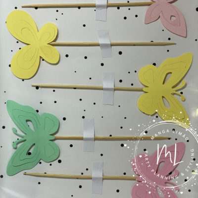 Pregadores decorativos em forma de borboletas coloridas em paus de madeira sobre fundo branco com pontos pretos.