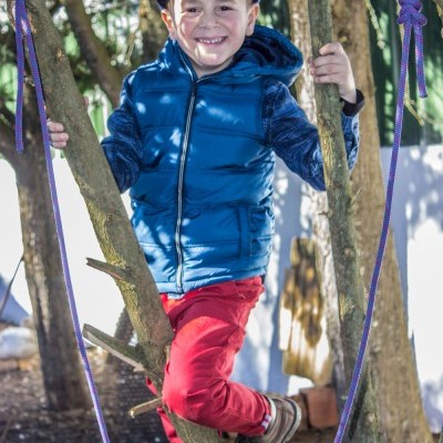 Criança com colete azul, calças vermelhas e boné azul pendurada numa corda roxa entre árvores