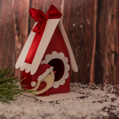 Casa de passarinho vermelha e branca com laço vermelho, raminho verde e neve falsa sobre superfície de madeira