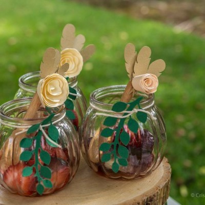 Quatro frascos de vidro com decoração de folhas e flores sobre um tronco de árvore em espaço ao ar livre