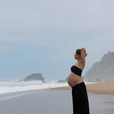 Mulher grávida de top e saia pretos na praia com mar e pedras ao fundo