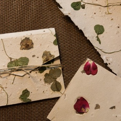 Envelopes e papel artesanal com folhas e pétalas secas sobre tecido castanho.
