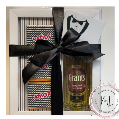Conjunto de presente com garrafa de whisky Grant's, duas barras de chocolate embaladas e cartão, envolto em laço preto dentro de caixa branca
