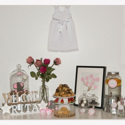 Mesa decorada com bolo, jarra, flores, placa WELCOME RITA e vestido branco pendurado na parede