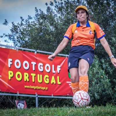 Mulher em uniforme de footgolf a controlar bola branca em campo junto a cartaz FOOTGOLF PORTUGAL