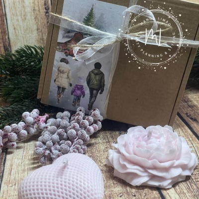 Caixa de cartão com fita branca e etiqueta decorativa, rodeada por bagas e velas rosa claro em superfície de madeira