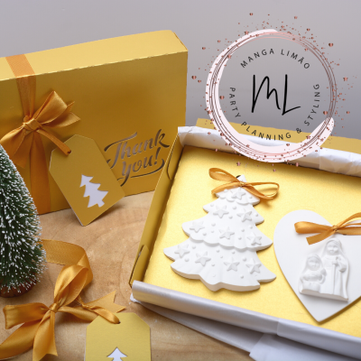 Caixa dourada com laço e etiquetas amarelas, ornamentos brancos em formato de árvore e coração, árvore decorativa verde e selo MANGA LIMÃO.