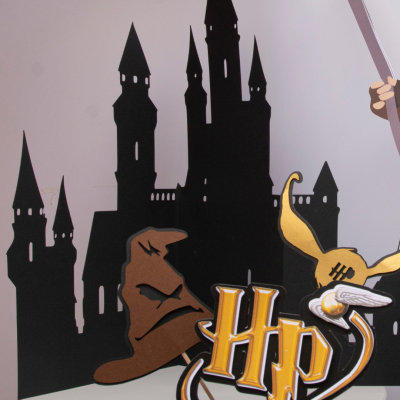 Ilustração de castelo preto com chapéu marrom, ‘HP’, Golden Snitch e varinha mágica