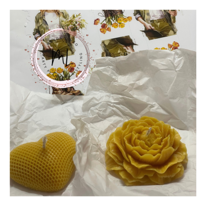 Duas velas amarelas de cera em formatos de coração e flor sobre papel branco com imagens ao fundo