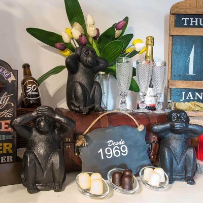 Três estatuetas de macacos pretos em poses diferentes, garrafa de champanhe, copos, calendário de madeira, bolo e arranjo floral.